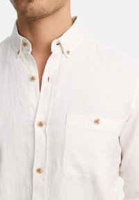 Camisa blanca de lino con cuello abotonado, que presenta botones de madera y un bolsillo en el pecho. La tela tiene un acabado texturizado.