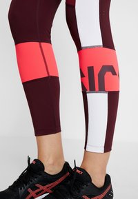 Viininpunaiset ja vaaleanpunaiset urheilutrikoot, joissa on värilohkoja, valkoisia yksityiskohtia ja logotunnus; sileä, joustava kangas. Mustat lenkkarit alla.