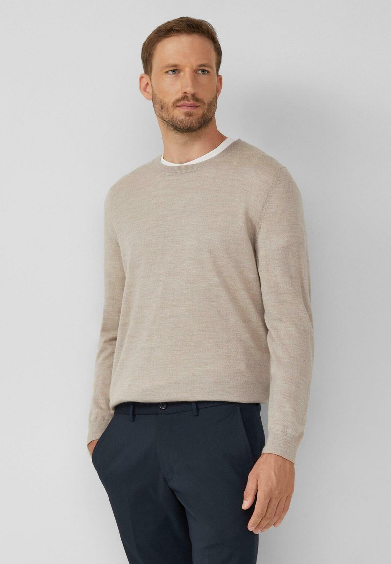 Jas-beżowy sweter z miękkiego materiału o klasycznym okrągłym dekolcie i długich rękawach, zestawiony z ciemnymi granatowymi spodniami.