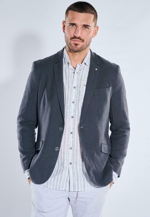 Man wearing dark blazer over light striped button-up shirt and white pants, standing with hands in pockets against plain background.