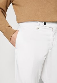 Pantaloni bianchi in tessuto liscio, con tasca frontale, chiusura a bottone nera e accessori in argento sulla cintura.