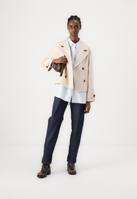 Giacca beige doppiopetto, camicia a righe azzurre, pantaloni in denim scuro e stivali alla caviglia marroni. Tessuto liscio con vestibilità su misura.