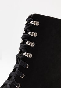 Botín negro de cordones que presenta un material similar al ante, ojales metálicos y cordones negros planos, con un diseño elegante y estructurado.