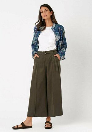 Woman wearing a white t-shirt, blue floral jacket, olive wide-leg cropped pants with belt, and dark brown flat sandals, standing with hands in pockets.
