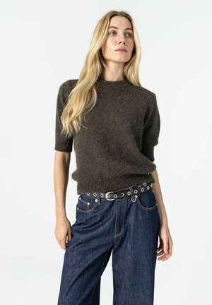 Blonde woman wearing a dark brown fuzzy short-sleeve sweater, blue jeans, and a black belt with silver eyelets, standing against a white background.