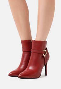 Stivali alla caviglia in pelle rossa con punta affusolata, tacco stiletto sottile e una cinghia decorativa con un accento in metallo dorato circolare.