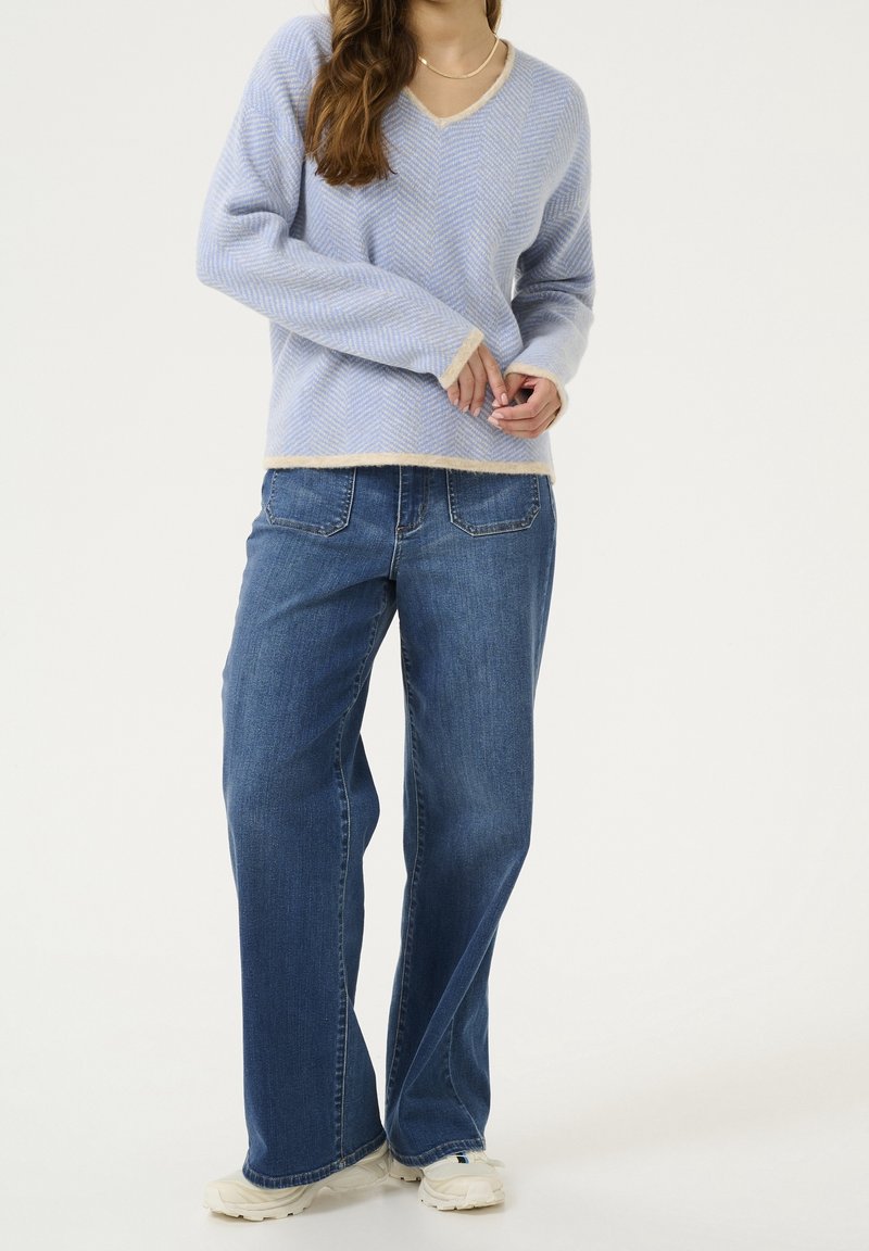 Femme portant un pull col V bleu clair, un jean bleu large et des baskets blanches, debout sur fond blanc.