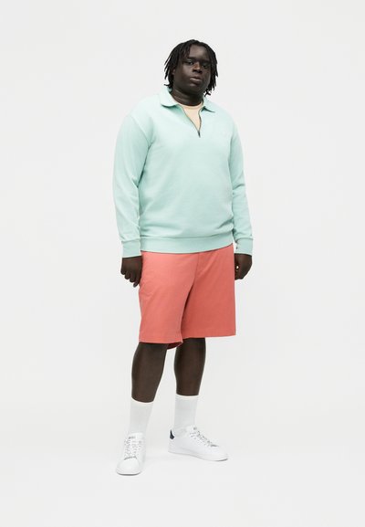 Moški stoji v svetlo turkizni jopiči s četrtinskim zadrgo, koralnordečih kratkih hlačah do kolen, belih nogavicah in belih supergah na enobarvnem ozadju.