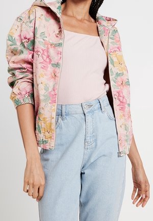 Frau trägt eine rosa florale Reißverschlussjacke über einem hellrosa gerippten Top und hoch taillierte hellblaue Jeans mit entspanntem Schnitt.