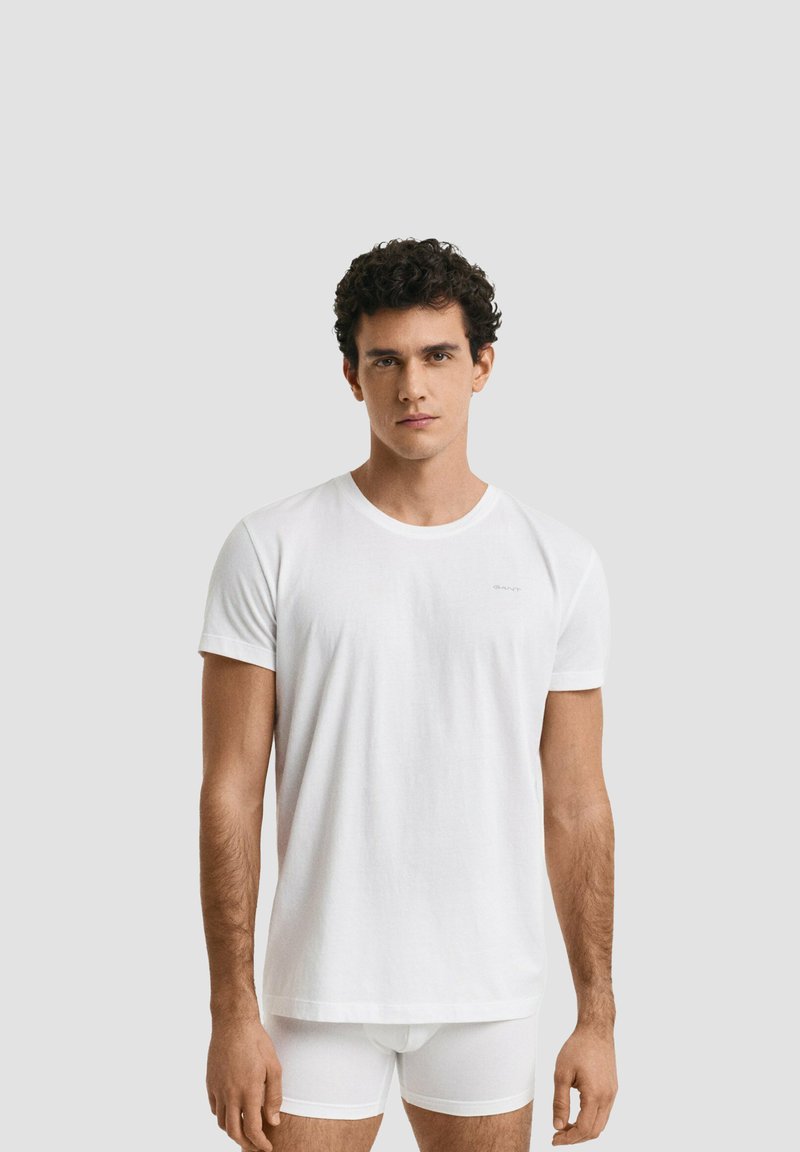 Giovane uomo con capelli ricci indossa una maglietta bianca semplice a maniche corte e boxer bianchi, in piedi davanti a uno sfondo grigio chiaro.