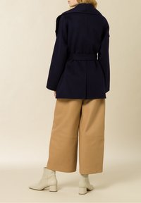 Abrigo de mezcla de lana color navy con solapas amplias, cintura ceñida con cinturón y hombros estructurados, combinado con culottes de cuero color tostado y botines de ante color crema.
