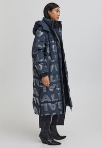 Abrigo largo acolchado, azul marino oscuro con grandes letras reflectantes, cierre de cremallera y capucha. Combinado con botines negros y vaqueros desgastados.