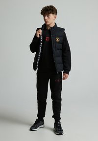 Giacca a puffo nera con un patch logo, indossata sopra una maglietta nera, abbinata a pantaloni neri e scarpe da ginnastica grigie, su uno sfondo neutro.