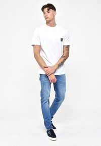 T-shirt bianca in cotone con taschino sul petto, abbinata a jeans di denim blu chiaro. Le calzature sono un paio di sneakers blu scuro. Look pulito e casual.