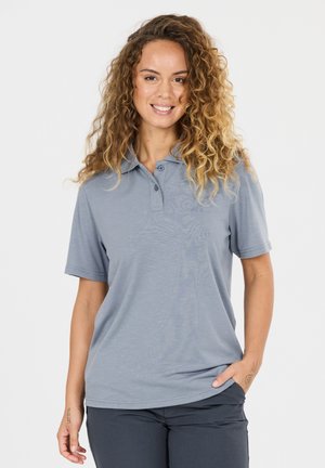 Frau mit lockigem Haar, die ein hellblaues kurzärmeliges Hemd mit Kragen und dunkle Hosen trägt, lächelt und hat eine Hand in der Tasche.