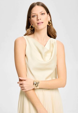 Mujer joven con cabello castaño claro lleva una blusa sin mangas color crema, falda plisada a juego, grandes pendientes de aro dorados y una pulsera gruesa dorada.