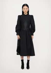 Zwarte jurk met een aansluitend leren lijfje, geplooide mouwen, een getailleerde taille met gespsdetails en een vloeiende rok. Accentaccessoires omvatten een tas.
