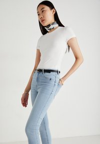 Λευκό εφαρμοστό t-shirt συνδυασμένο με ανοιχτό μπλε στενό τζιν. Το μοντέλο φοράει μία μαύρη δερμάτινη ζώνη και ένα μαντήλι με ασπρόμαυρο σχέδιο.