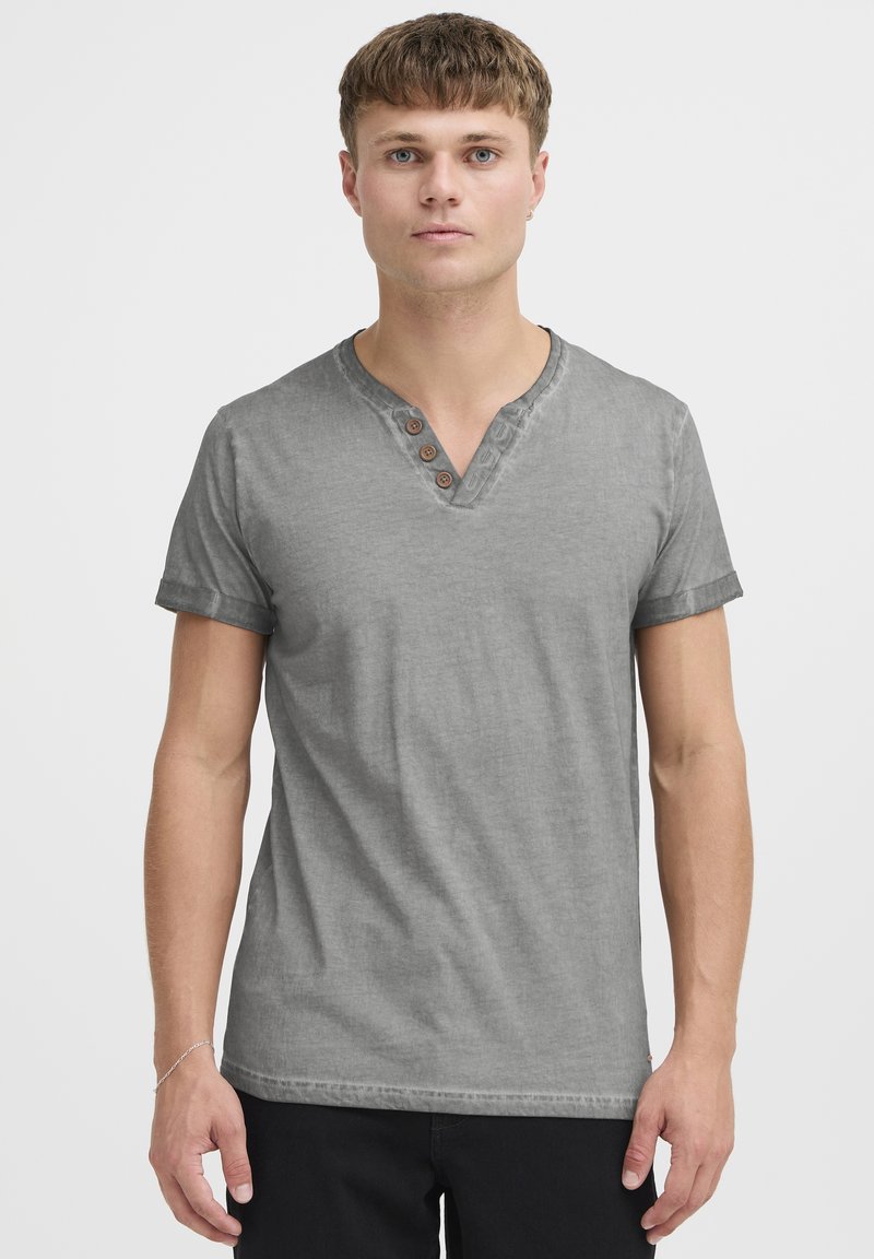 Camiseta gris de manga corta con escote en V y tres botones de madera, con una textura suave y mangas enrolladas, llevada por una persona.