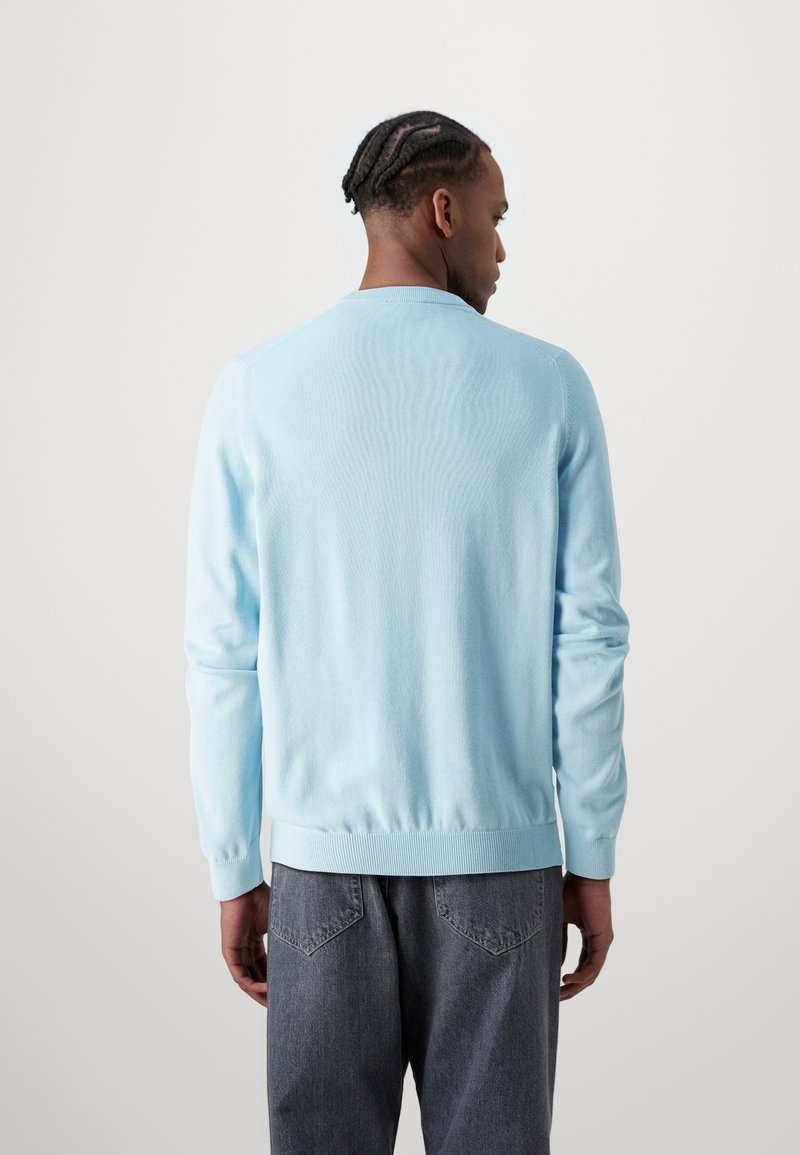 Pull en maille bleu clair avec un col rond et un ourlet côtelé, comportant des manches longues et une texture lisse. Porté sur un pantalon en denim gris.