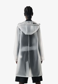 Person with braided hair wearing a semi-transparent white raincoat with a hood, standing facing away against a plain background.