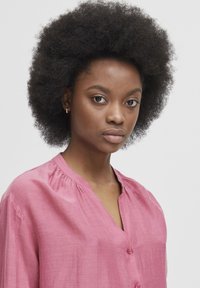 Blusa rosa dalla vestibilità ampia realizzata in tessuto leggero, con scollatura a V, chiusura con bottoni e dettagli arricciati attorno al colletto.
