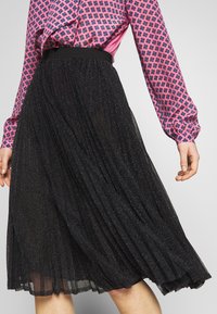 Musta pleitetty midihame, jossa on kimaltava pinta, joustava vyötärö ja lentoisa muoto. Kuvassa näkyy kuviollinen vaaleanpunainen paita.