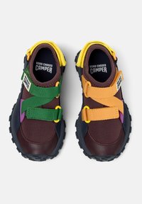 Ett par barn sneakers i mörkbrunt med gula kragar, gröna och orangea korsremmar, svarta sulor och texten "Bobo Choses Camper" inuti.