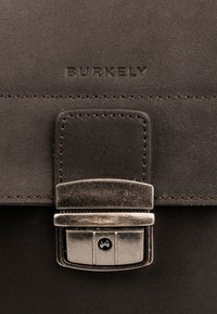 Bruine leren tas met een gladde textuur, voorzien van een metalen sluiting en het gegraveerde "BURKELY" logo aan de voorkant. Minimalistisch ontwerp.