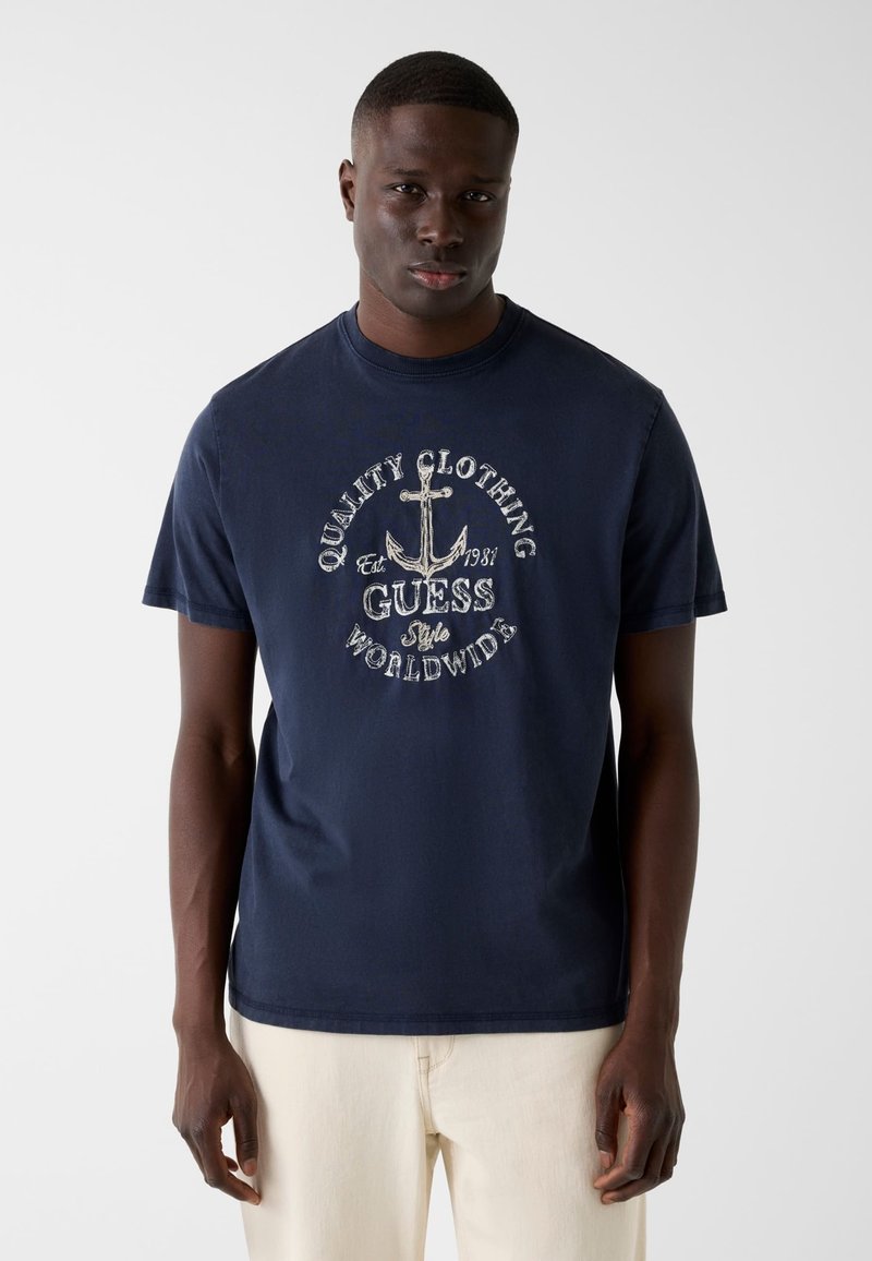 Άνδρας που φοράει μπλε σκούρο T-shirt με λευκό γραφικό άγκυρας και το κείμενο "Quality Clothing Guess Style Worldwide" σε συνδυασμό με ανοιχτό μπεζ παντελόνι.