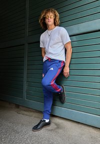 Dvoubarevný sportovní outfit složený ze šedého trička a modrých joggingových kalhot s červenými bočními pruhy a logem Adidas. Černé tenisky dokončují vzhled.
