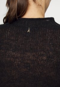 Personne aux cheveux bruns portant un pull en maille noir avec une petite broche dorée en forme d’insecte sur le haut du dos près de l’encolure.