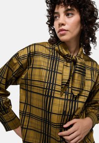 Chemise boutonnée en jaune moutarde avec un motif à carreaux noirs. Présente un col à nœud papillon et des manches longues. Tissu lisse et léger.