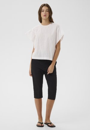 Vrouw draagt een witte top met korte mouwen, zwarte knielange broek en zwarte platte sandalen, staand tegen een effen achtergrond.