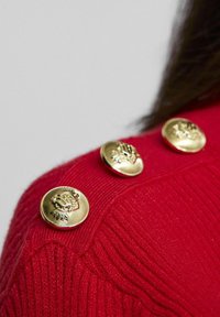 Röd stickad tröja med tre gyllene knappar med intrikata mönster på axeln. Texturen är ribbad, vilket framhäver dess detaljerade struktur.