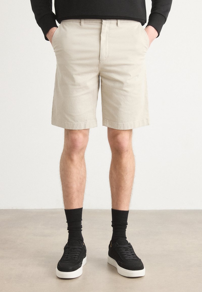 Beige cotton shorts with a straight cut, side pockets, and a smooth texture, paired with black sneakers and crew socks.