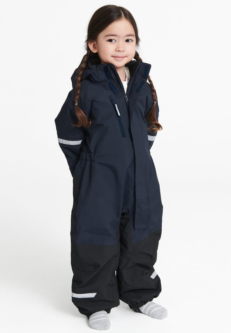 Laivastonsininen vedenpitävä haalari mustilla yksityiskohdilla; varustettu hupulla, heijastinliuskoilla ja joustavilla hihansuilla. Suunniteltu kylmälle säälle.