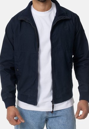 Mann trägt marineblaue Jacke mit Reißverschluss über weißem T-Shirt und hellblauen Jeans, steht mit herunterhängenden Händen vor schlichten Hintergrund.