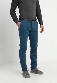 Homme portant un pantalon bleu ajusté, une chemise grise texturée à manches longues, une ceinture marron et des baskets grises, debout avec une main dans la poche.