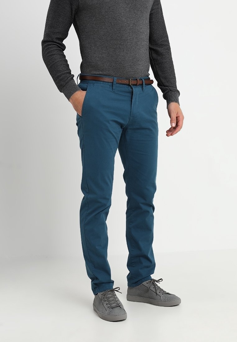 Homme portant un pantalon bleu ajusté, une chemise grise texturée à manches longues, une ceinture marron et des baskets grises, debout avec une main dans la poche.