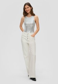 Zilveren mouwloze top en hoge witte broek met een knoopdetail. De broek heeft zijzakken en een rechte pijp.