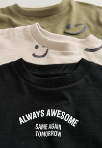 Trois t-shirts à col rond empilés : vert olive, beige avec un graphique de sourire, et noir avec un texte blanc imprimé portant la mention "TOUJOURS GÉNIAL, MÊME CHOSE DEMAIN."