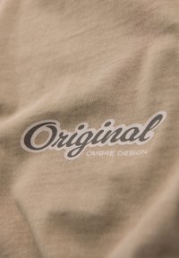 Béžová látka s hladkým povrchem, která nese výrazné černobílé logo se slovy "Originální OMBRE DESIGN." Jednoduchý a moderní design.