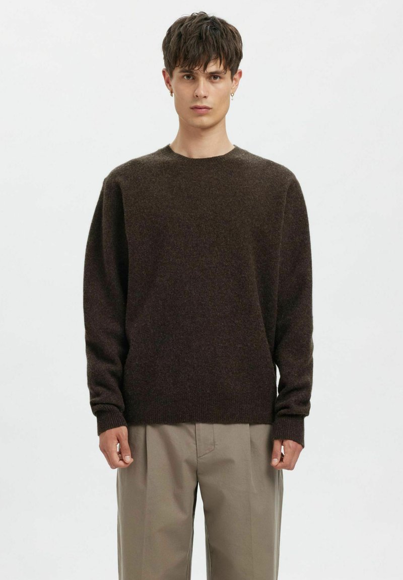 Rjavi pleteni pulover z okrolim izrezom, sproščenega kroja in dolgimi rokavi. Mehka tekstura, rebrasti manšeti in spodnji rob, kombiniran s bež hlačami.