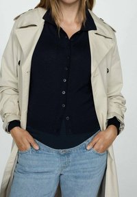 Beige trenchcoat over een marineblauwe button-up sweater, gecombineerd met lichtblauwe jeans. De jas heeft een klassiek kraag en gedetailleerde knopen.