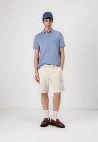 Polo shirt azzurro chiaro, pantaloni cargo beige con tasche laterali, mocassini marroni, calze bianche e un cappellino blu. Design semplice e texture morbida.