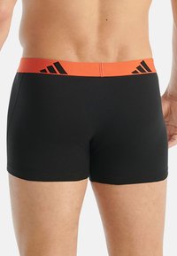 Černé plavky s oranžovým elastickým pasem a černými pruhy adidas. Materiál je hladký a přiléhavý, vhodný ke koupání.