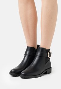 Bottines en cuir noir avec une semelle plate, fermeture éclair sur le côté et une sangle décorative dotée d'une boucle métallique ronde.