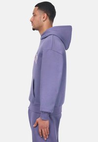 Sudadera morada con bolsillo frontal, puños y dobladillo acanalados. Tejido suave y texturizado que presenta un logo en el pecho. Capucha completa con cordones.