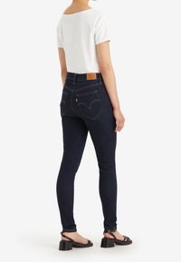 Jeans de mezclilla oscura con cintura alta, con bolsillos traseros sutilmente curvados y un parche de cuero marrón claro en la cintura. Combinados con una camiseta blanca.